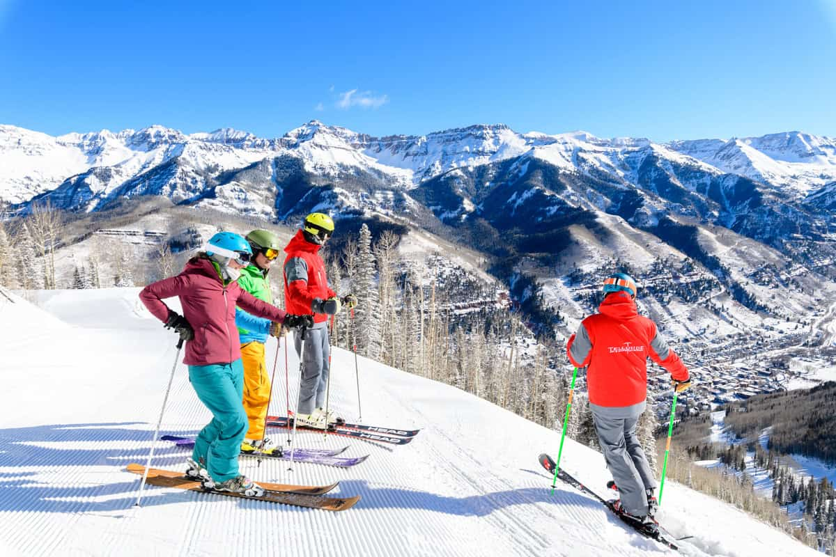 Four skiers in colorful outfits stand on a snowy slope overlooking a mountainous landscape under a clear blue sky.