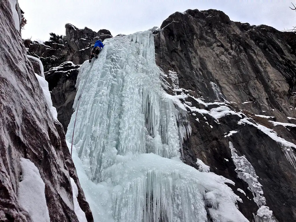 In a scene reminiscent of a guide to the best waterfalls, a climber in a blue jacket ascends a steep, frozen cascade on the rugged cliffside. Snow and ice blanket the surroundings, creating an awe-inspiring spectacle.