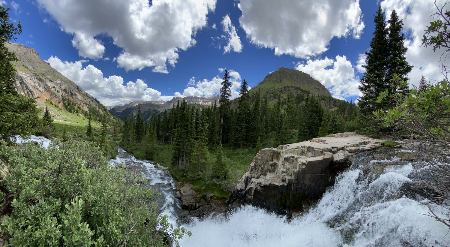 A scenic view of a waterfall flowing through a lush green forest with mountains and a partly cloudy blue sky in the background.