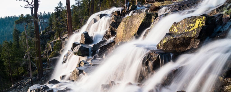 Water cascades over rocks in a forest setting, with blurred motion conveying a sense of flow and movement. Trees surround the waterfall, creating a natural, serene environment.