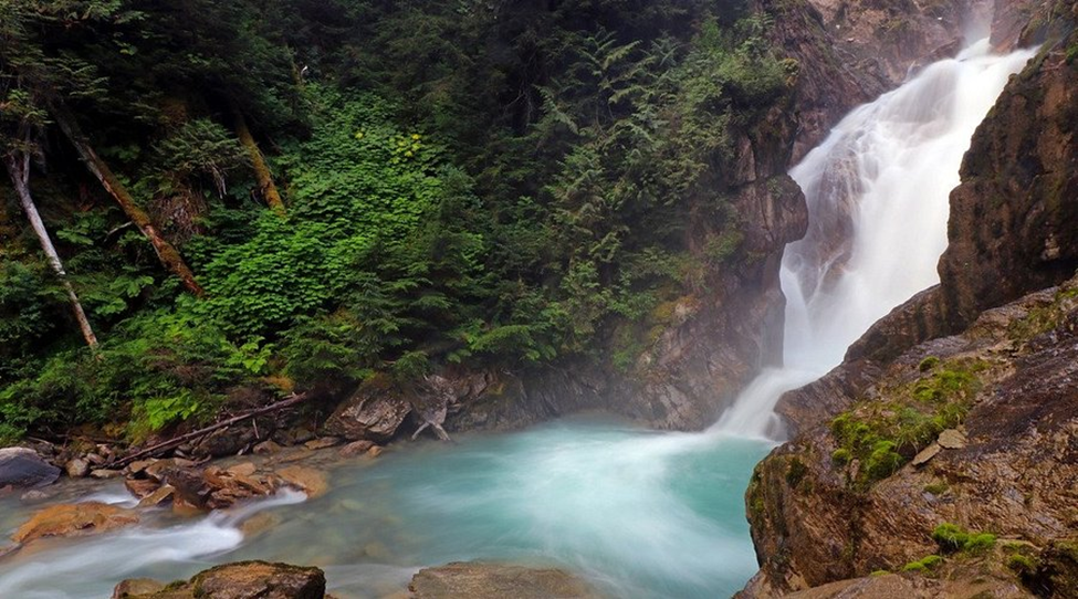 Waterfall cascading down rocks into a clear, teal pool surrounded by lush green foliage and rocky terrain.