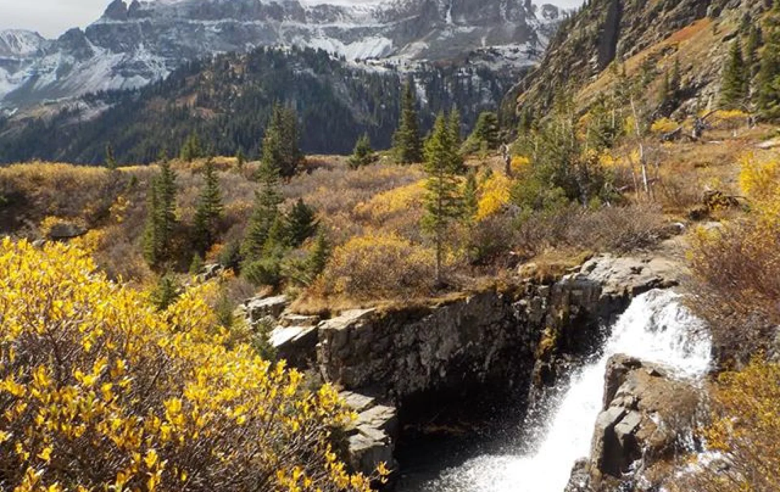 A small waterfall flows over rocks surrounded by autumn foliage and pine trees, with snow-capped mountains in the background.