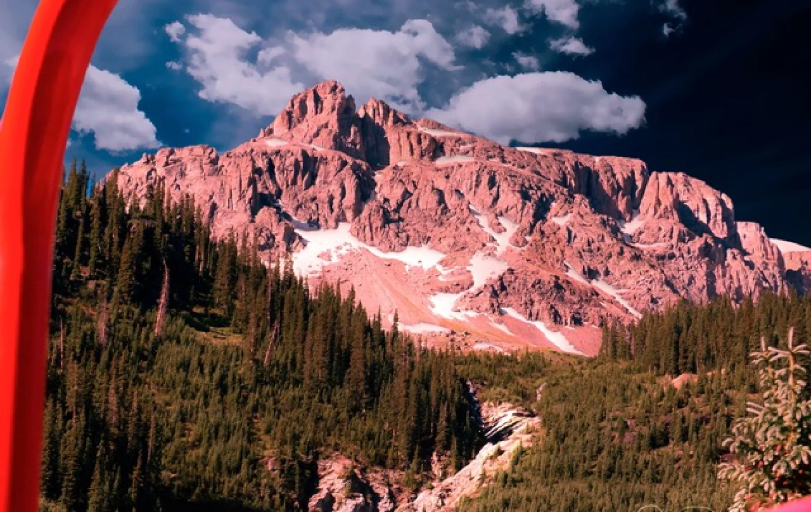 A mountain with patches of snow under a cloudy sky, surrounded by evergreen trees. A red arch is visible on the left side of the image.