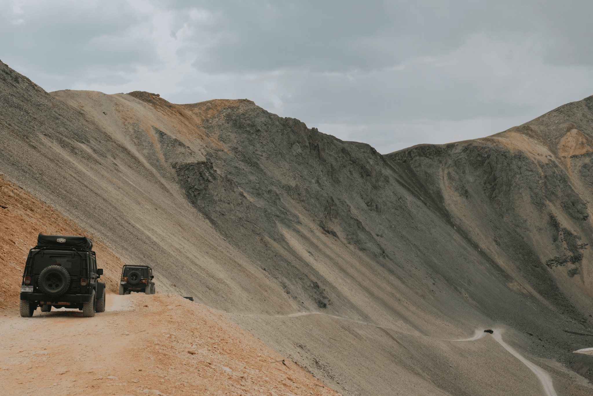 Two off-road vehicles drive on a narrow dirt trail along a steep mountainside under a cloudy sky.