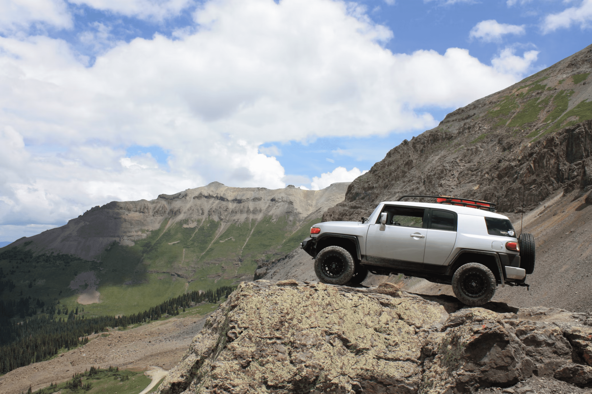 A white SUV with a roof rack parked on a rocky cliff with mountains and a cloudy sky in the background.