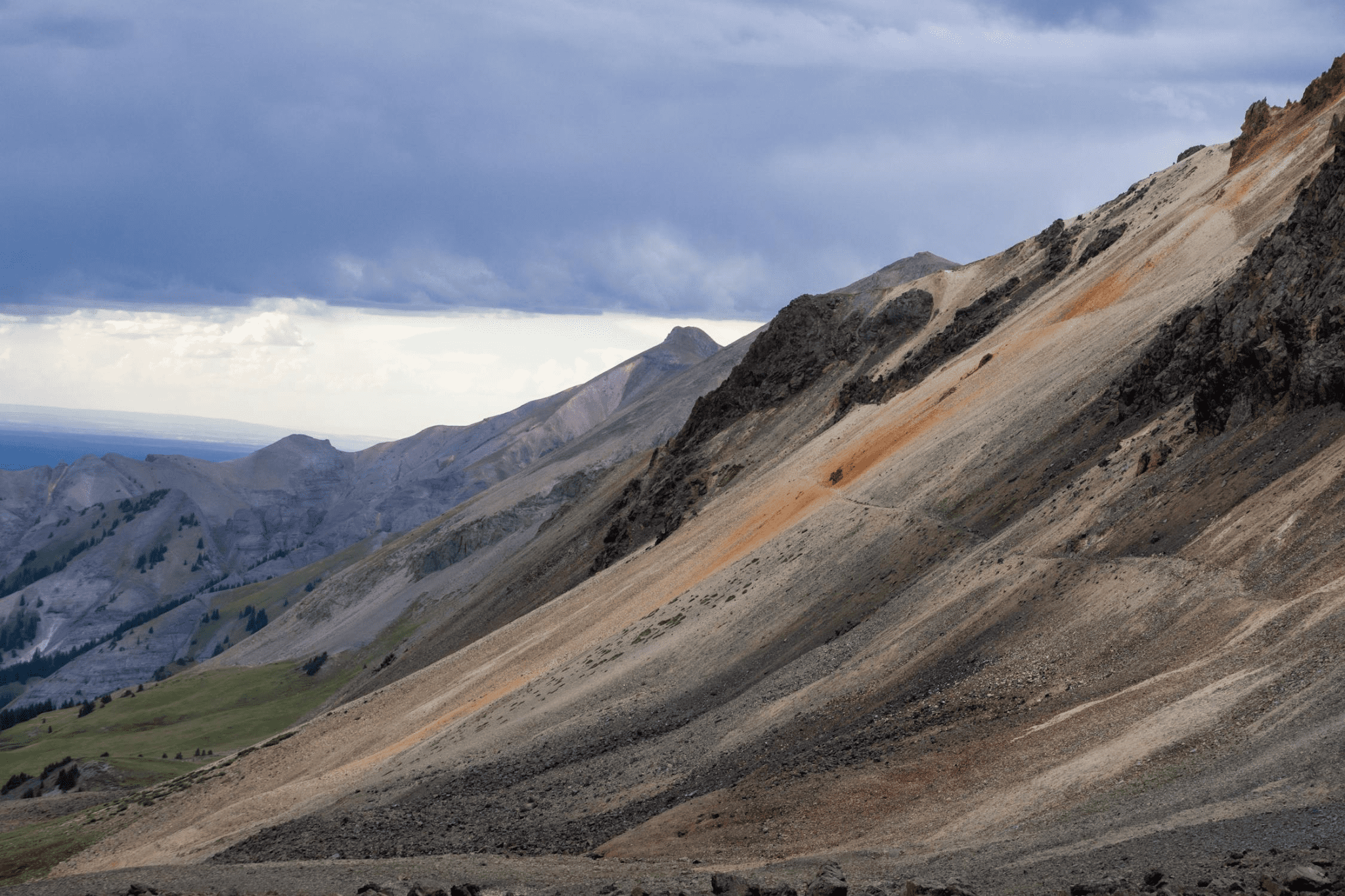 Mountain landscape with rocky slopes under a cloudy sky. Vegetation is sparse with varying hues of brown, gray, and green.