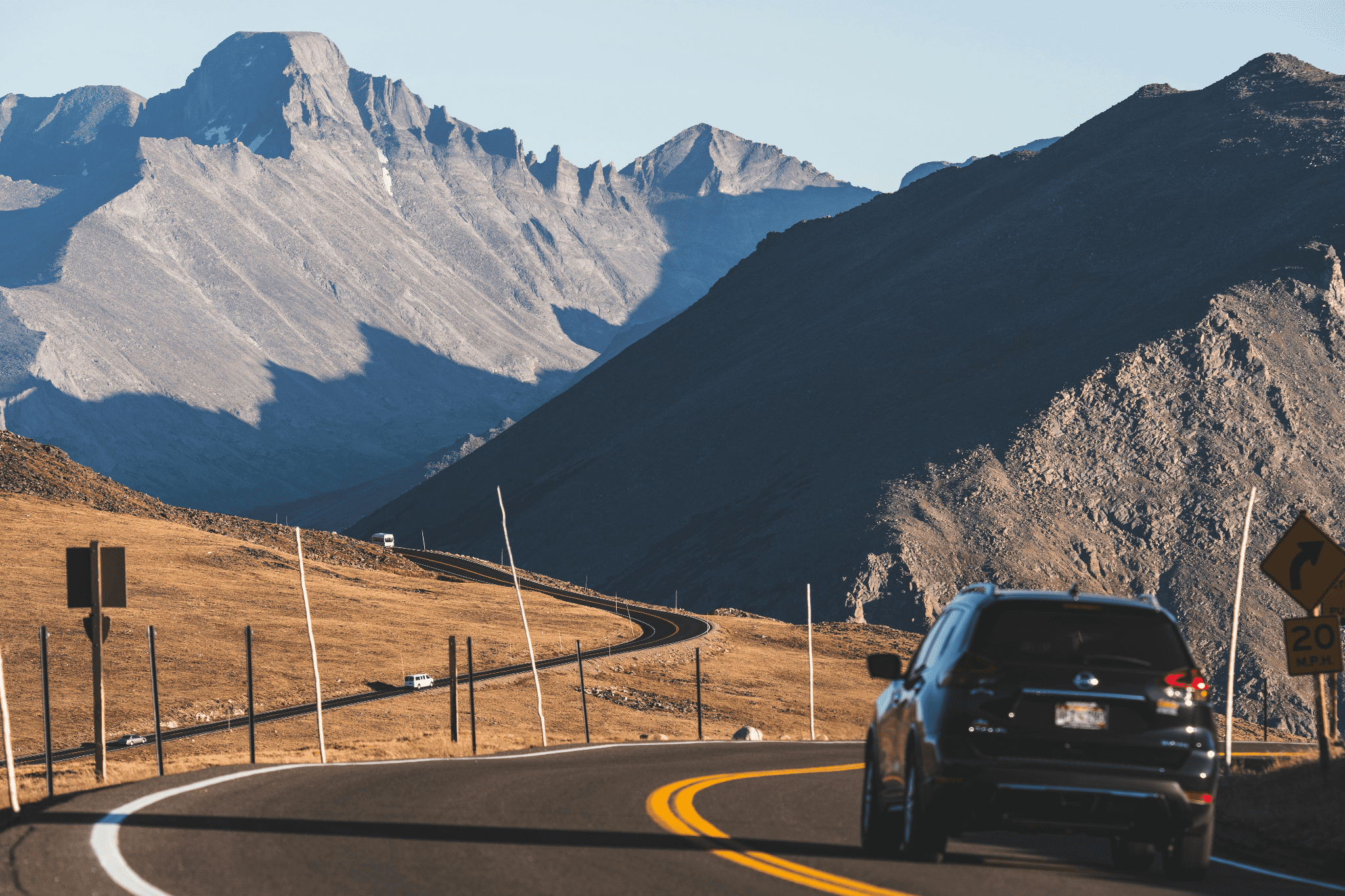 A car travels on a winding mountain road under clear skies, with rugged peaks in the background.