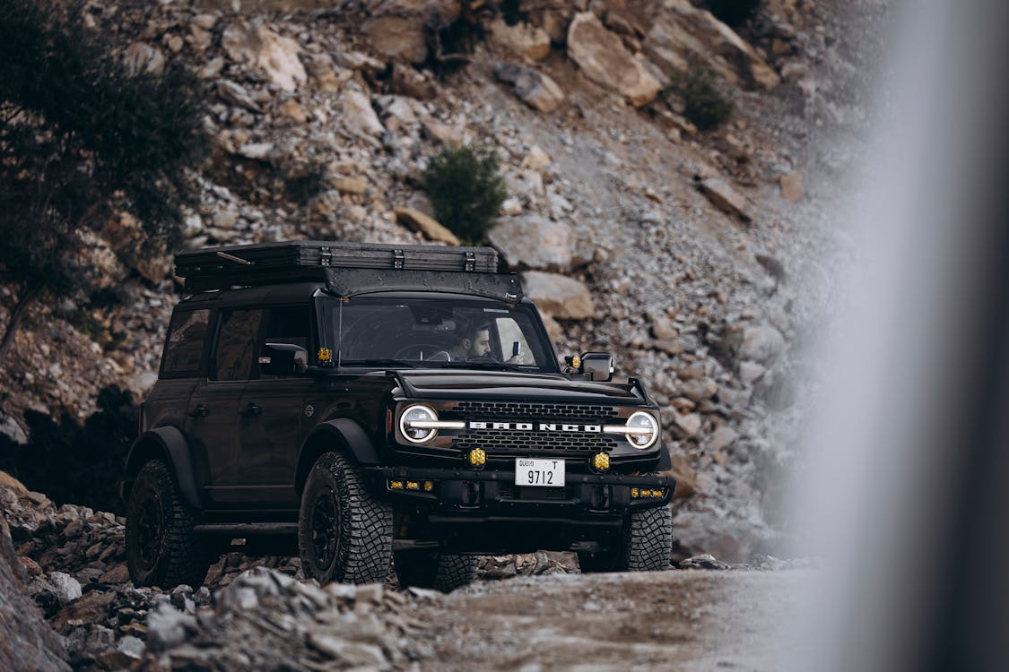 Black off-road SUV parked on rocky terrain, equipped with large tires and a roof rack, surrounded by rugged cliffs.