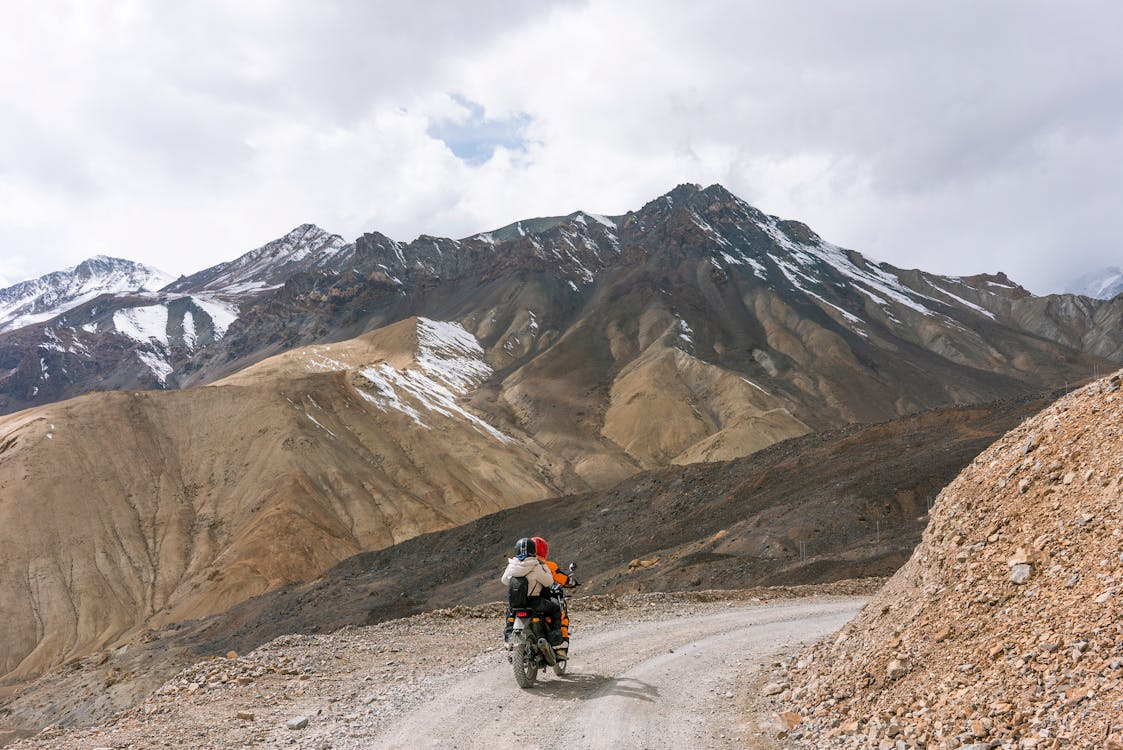 Motorcyclist riding on a gravel road through rugged mountains with patches of snow under a cloudy sky.