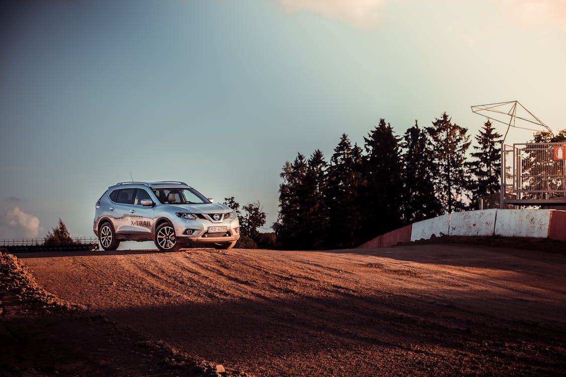 Silver SUV driving on a dirt road near a fenced area, with trees and a clear sky in the background.