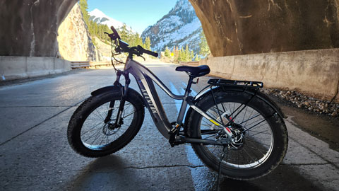 Electric bicycle parked in a mountain tunnel, with snow-capped peaks and pine trees visible in the background.