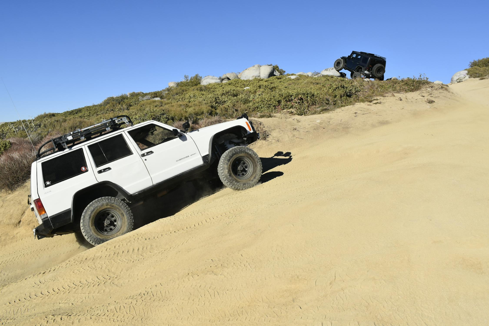 A white off-road SUV climbs a sandy hill while a second dark SUV is visible further ahead on the slope under a clear blue sky.