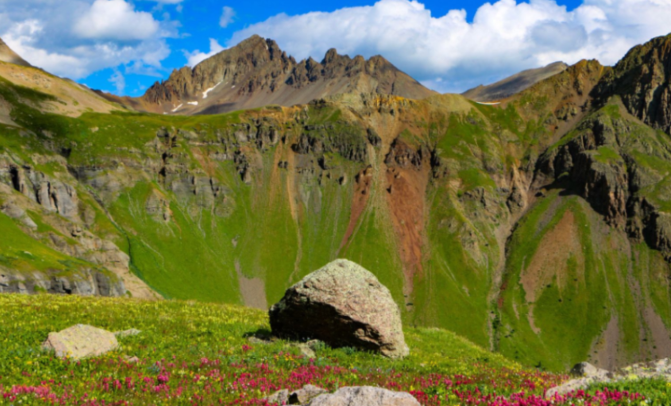 Rocky mountain landscape with green hills, patches of wildflowers in the foreground, and jagged peaks under a partly cloudy sky.