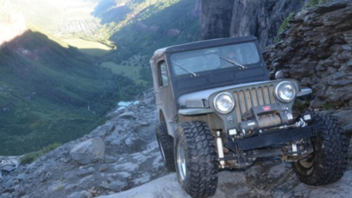A jeep driving on a steep, rocky mountain trail with a valley and distant landscape visible below.
