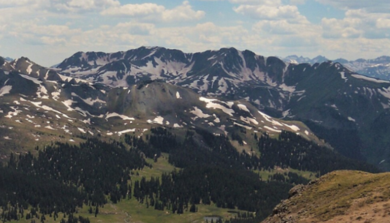 Mountain range with patches of snow, scattered trees, and grassy areas under a partly cloudy sky.