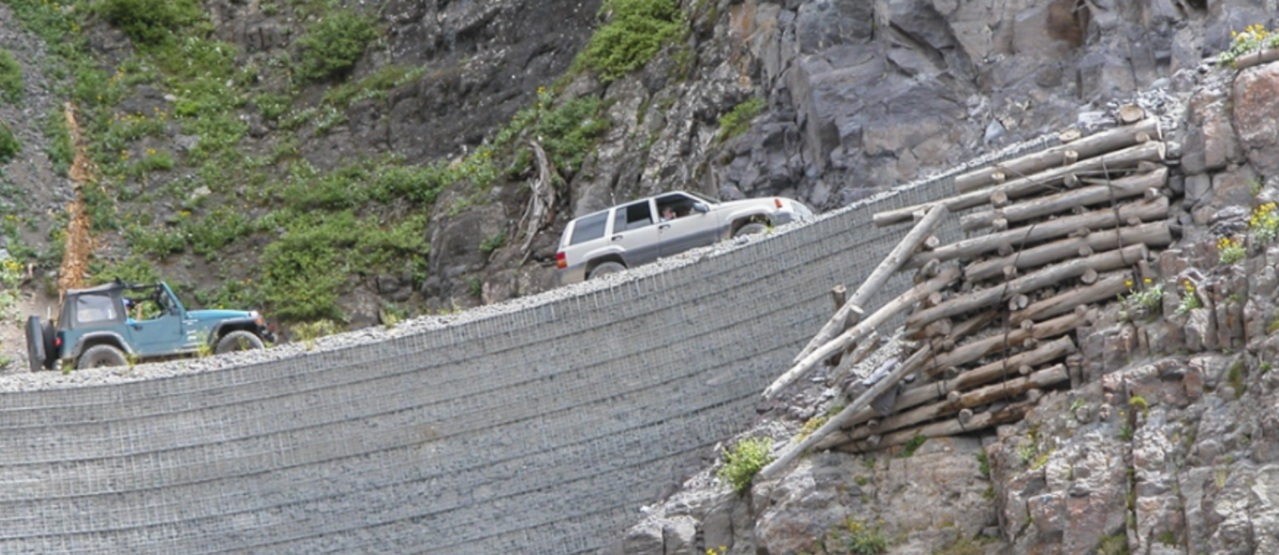 Two off-road vehicles drive on a narrow gravel road supported by a log and rock retaining wall along a rocky, steep mountainside.
