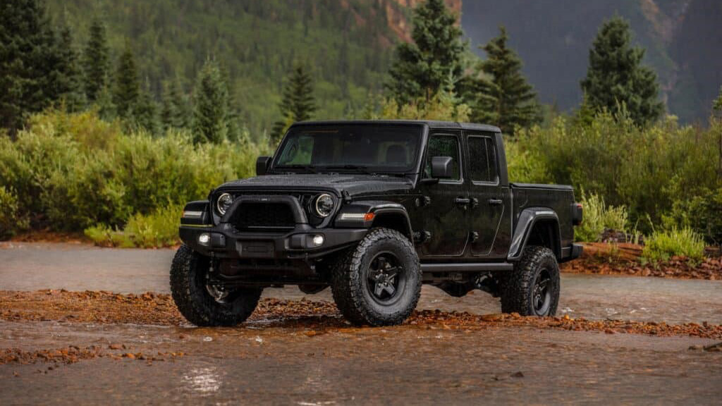 A black Jeep Gladiator pickup truck is parked on a muddy, rocky area with green trees and misty mountains in the background.