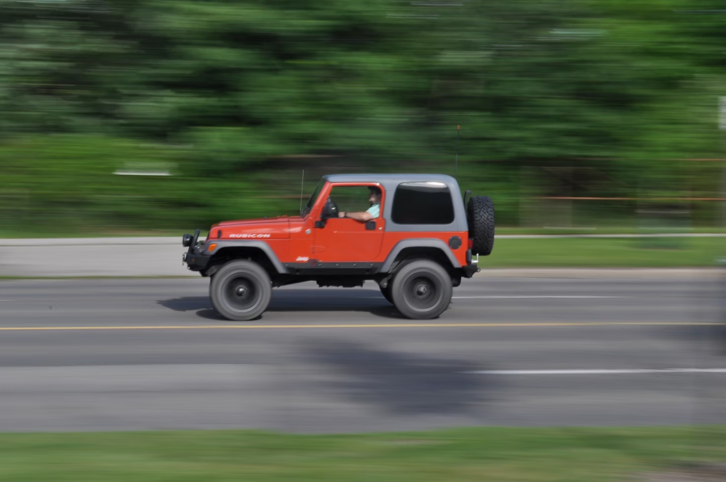 A red off-road vehicle drives along a street with blurred green trees and grass in the background, indicating motion.