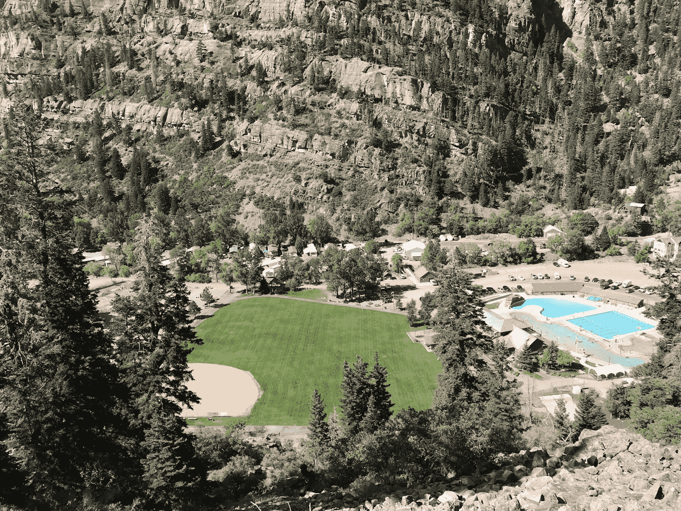 Aerial view of a green athletic field and a large outdoor swimming pool surrounded by trees and rocky hills.