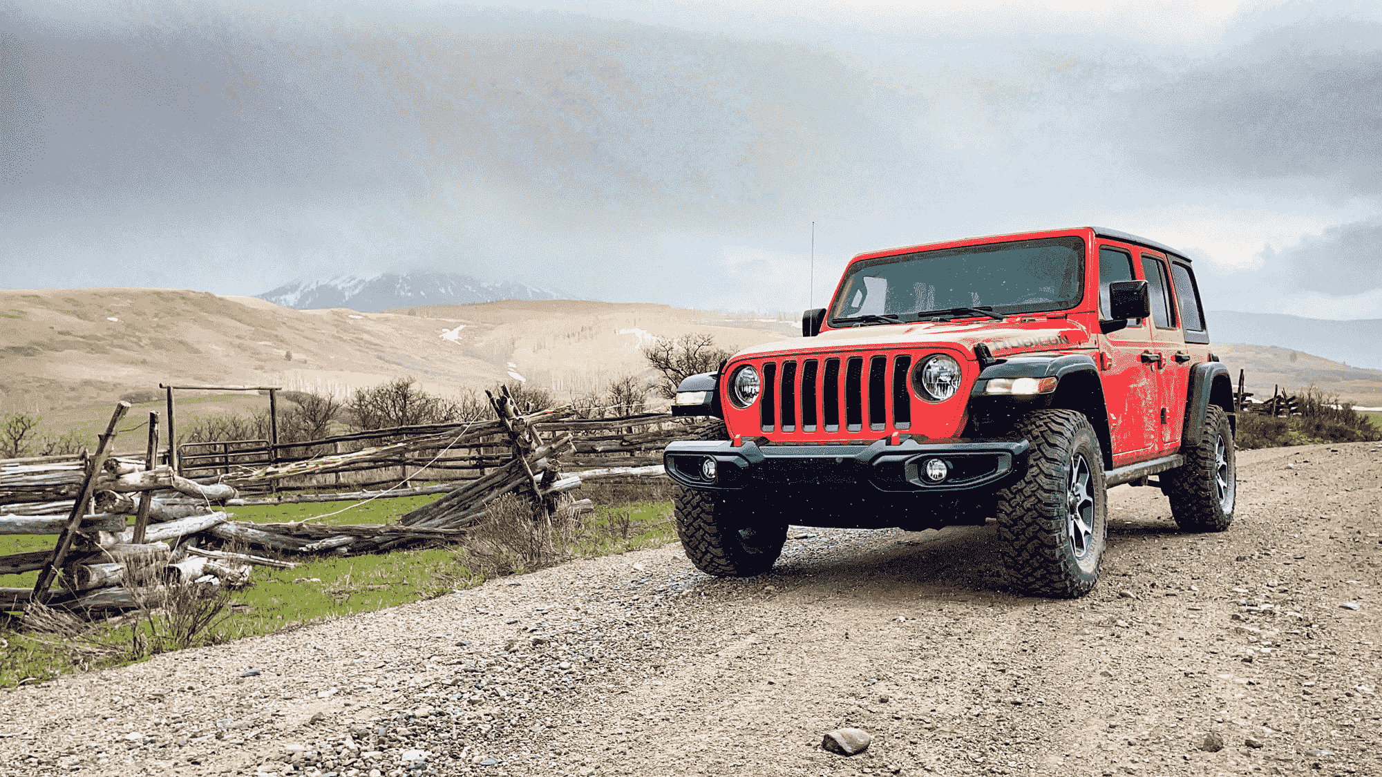 A red Jeep Wrangler is parked on a gravel road next to a wooden fence with hills and cloudy skies in the background.