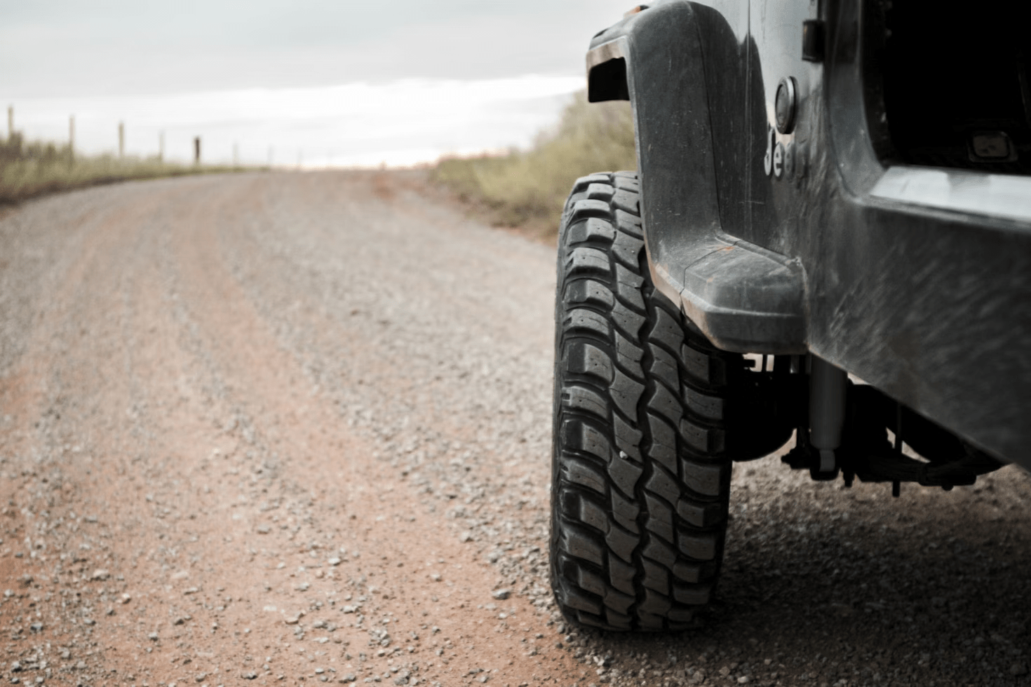 Close-up of a large off-road tire on a vehicle parked on a rural gravel road, with fields and a cloudy sky in the background.