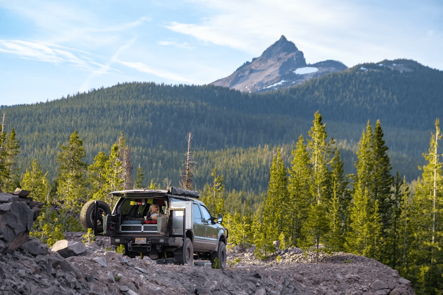 A pickup truck parked on a rocky trail in a forested area with a mountain peak in the background under a clear sky.