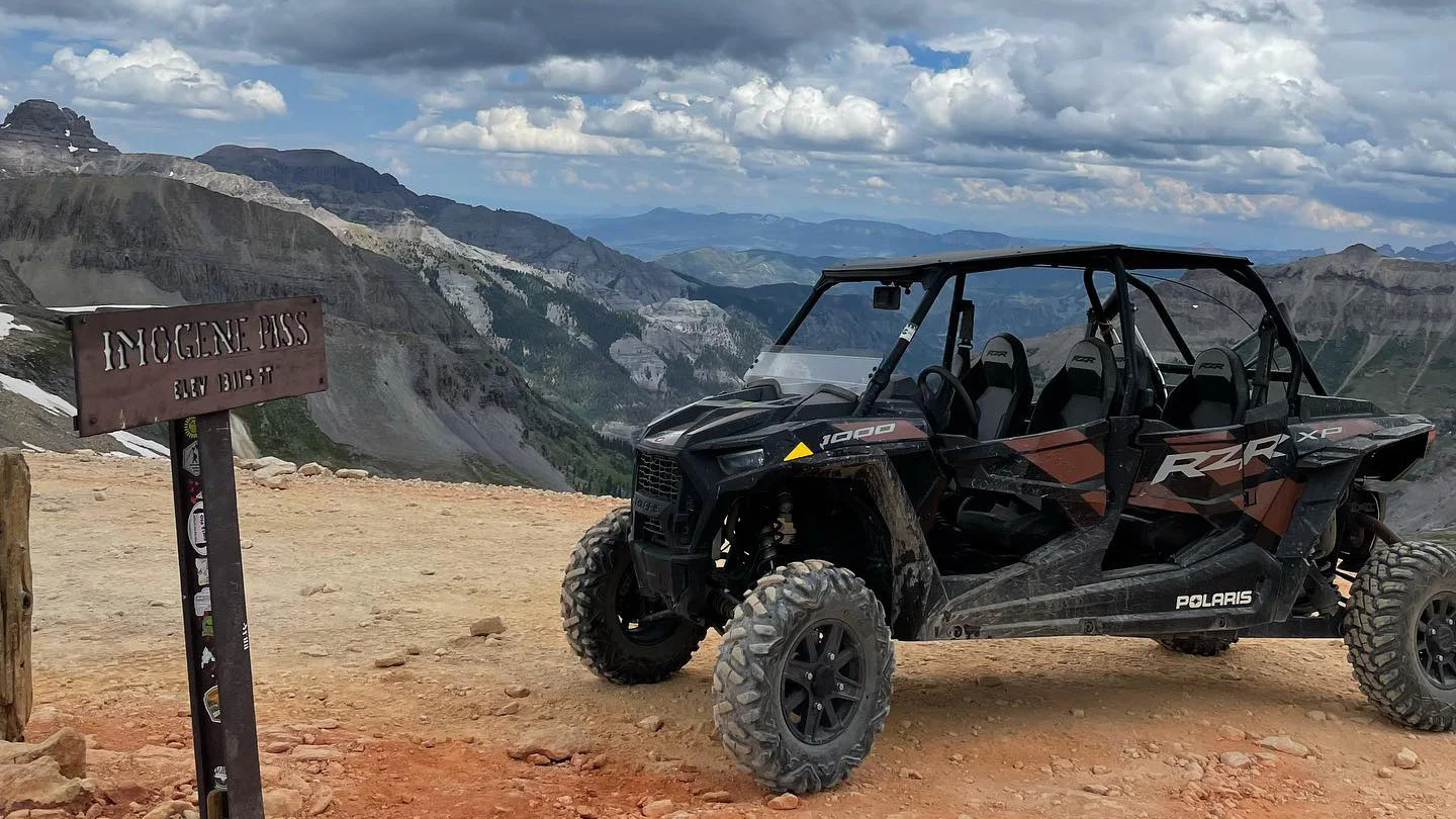 A Polaris RZR XP off-road vehicle is parked on a rocky mountain pass, with a wooden sign reading "Imogene Pass, Alt 13,114 ft" on the left and a scenic view of mountains and clouds in the background. It's an ideal spot for those looking to explore rugged terrains through off-road vehicle rentals.
